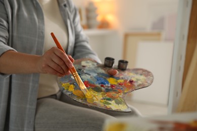 Photo of Woman with brush using palette near easel indoors, closeup