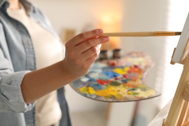 Photo of Woman holding brush and palette painting on easel with canvas indoors, closeup