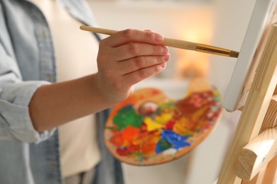 Photo of Woman holding brush and palette painting on easel with canvas indoors, closeup
