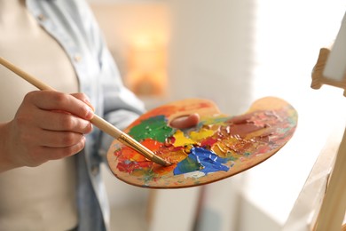 Photo of Woman mixing paints on palette with brush near easel indoors, closeup