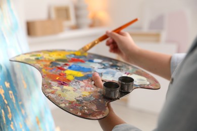 Photo of Woman mixing paints on palette with brush near easel indoors, closeup