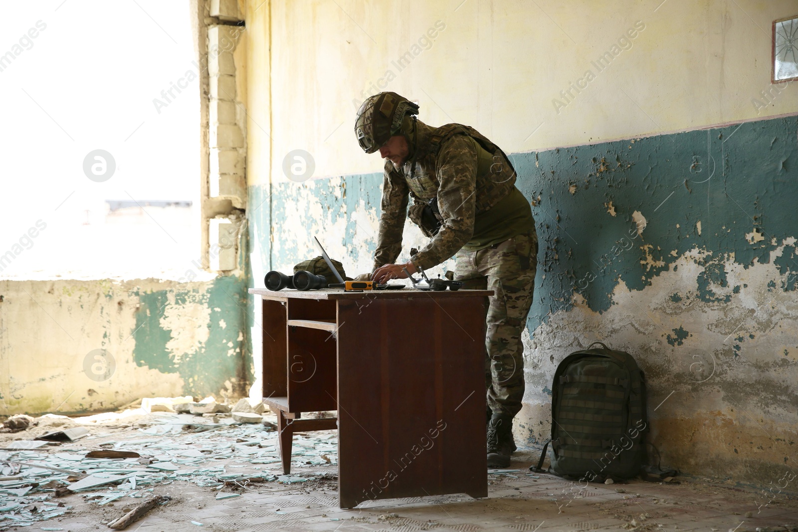 Photo of Military mission. Soldier in uniform using laptop at table inside abandoned building