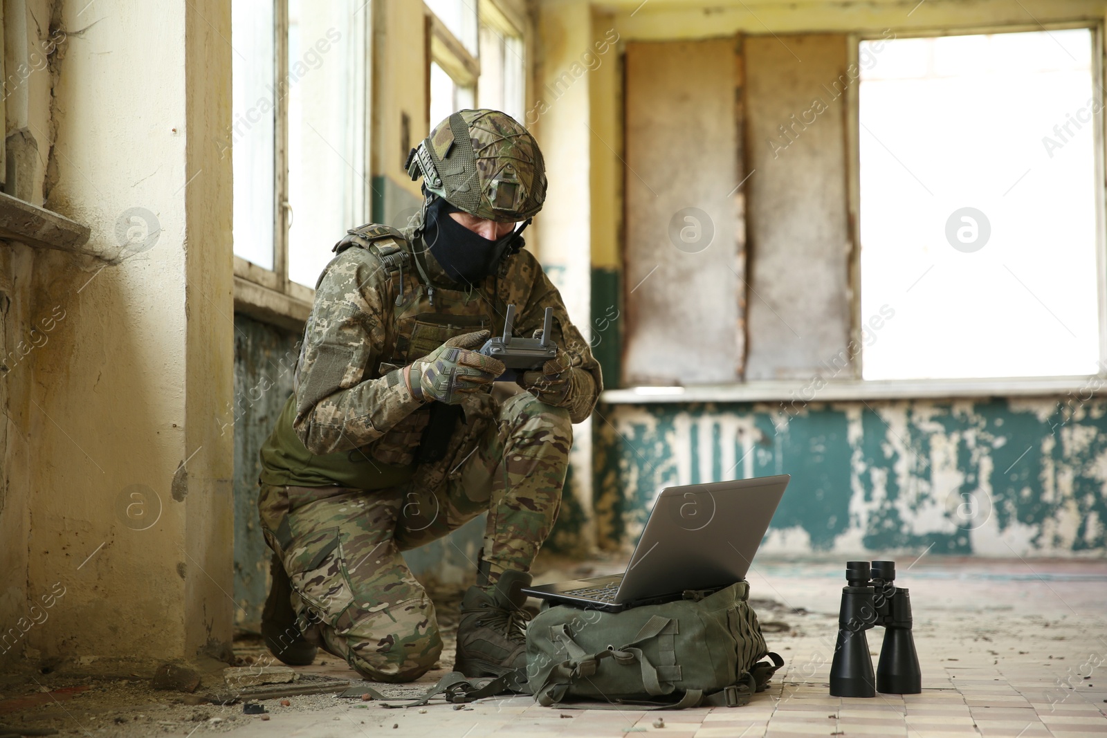 Photo of Military mission. Soldier in uniform using laptop and binoculars inside abandoned building