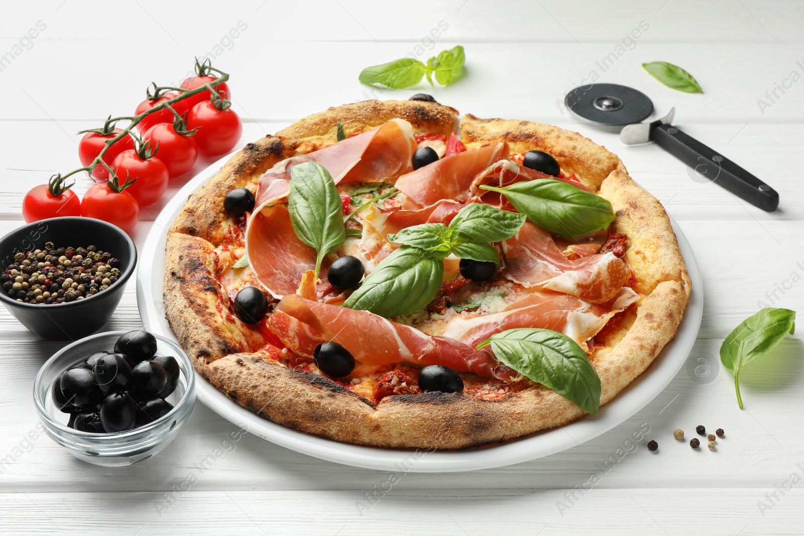 Photo of Tasty pizza with cured ham, olives, tomatoes and basil on white wooden table