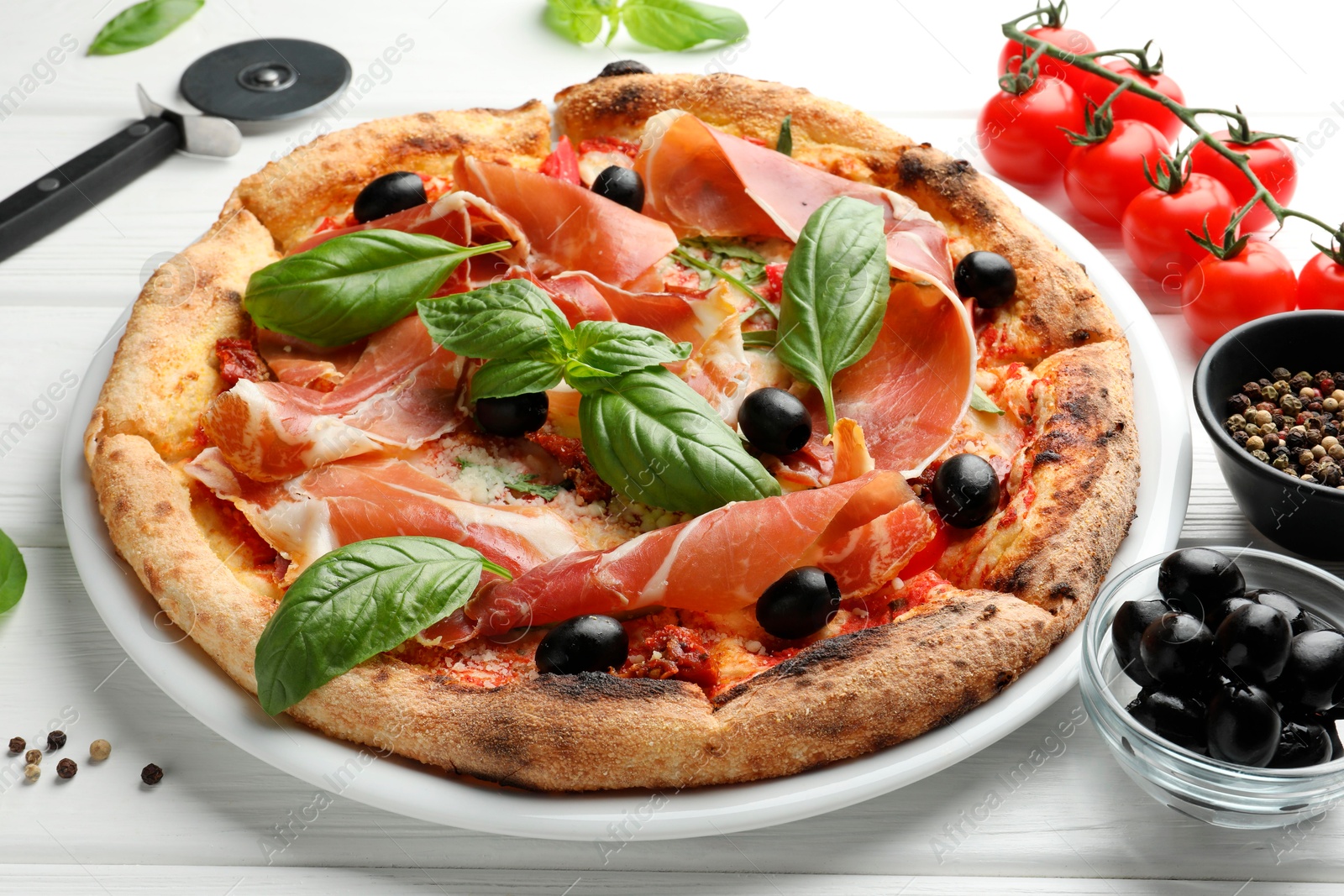 Photo of Tasty pizza with cured ham, olives, tomatoes and basil on white wooden table