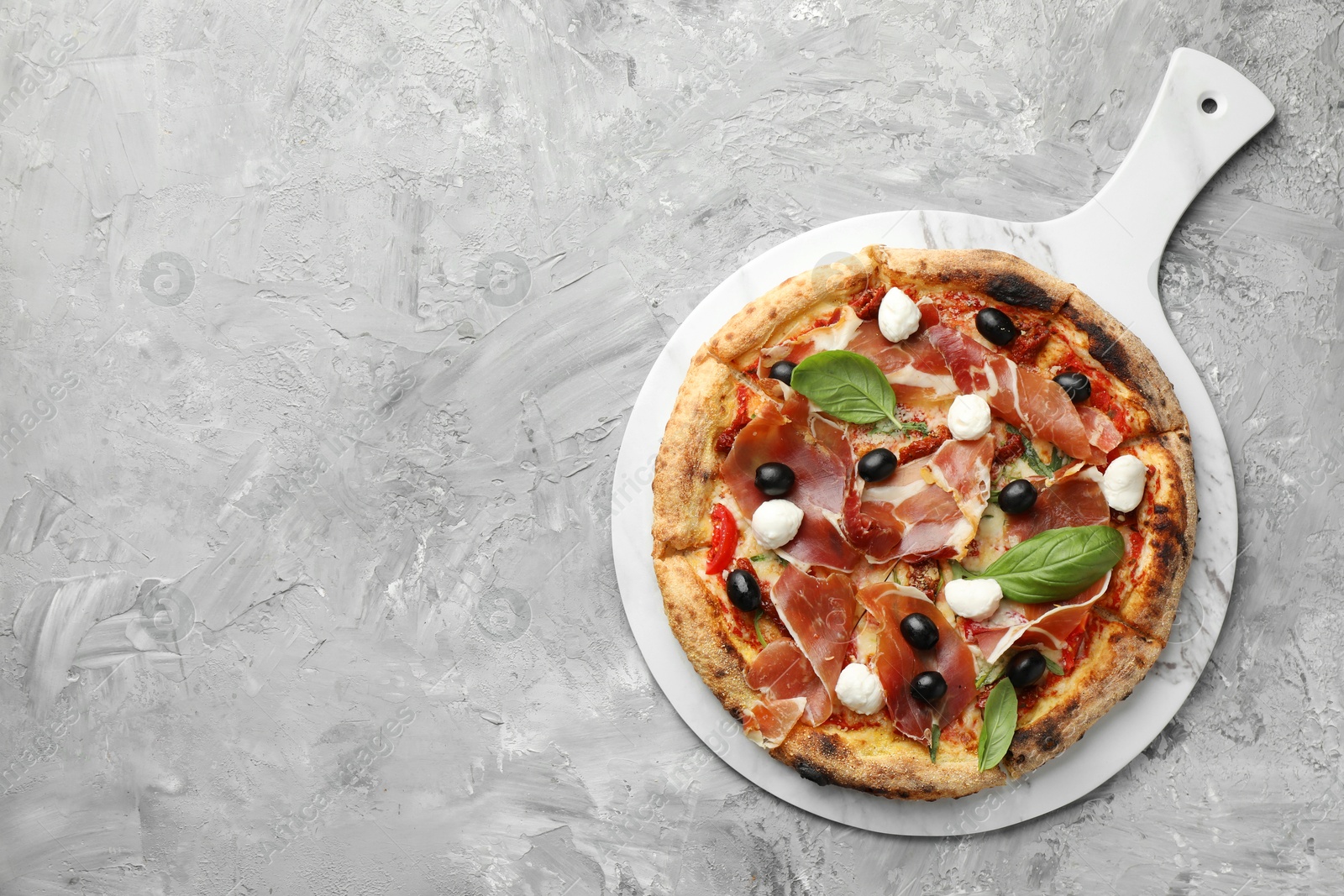 Photo of Tasty pizza with cured ham, olives, sun-dried tomato and basil on gray textured table, top view. Space for text