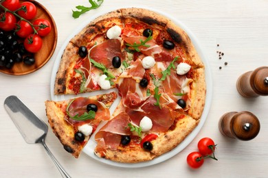 Tasty pizza with cured ham, olives, mozzarella cheese, tomatoes and arugula on light wooden table, flat lay