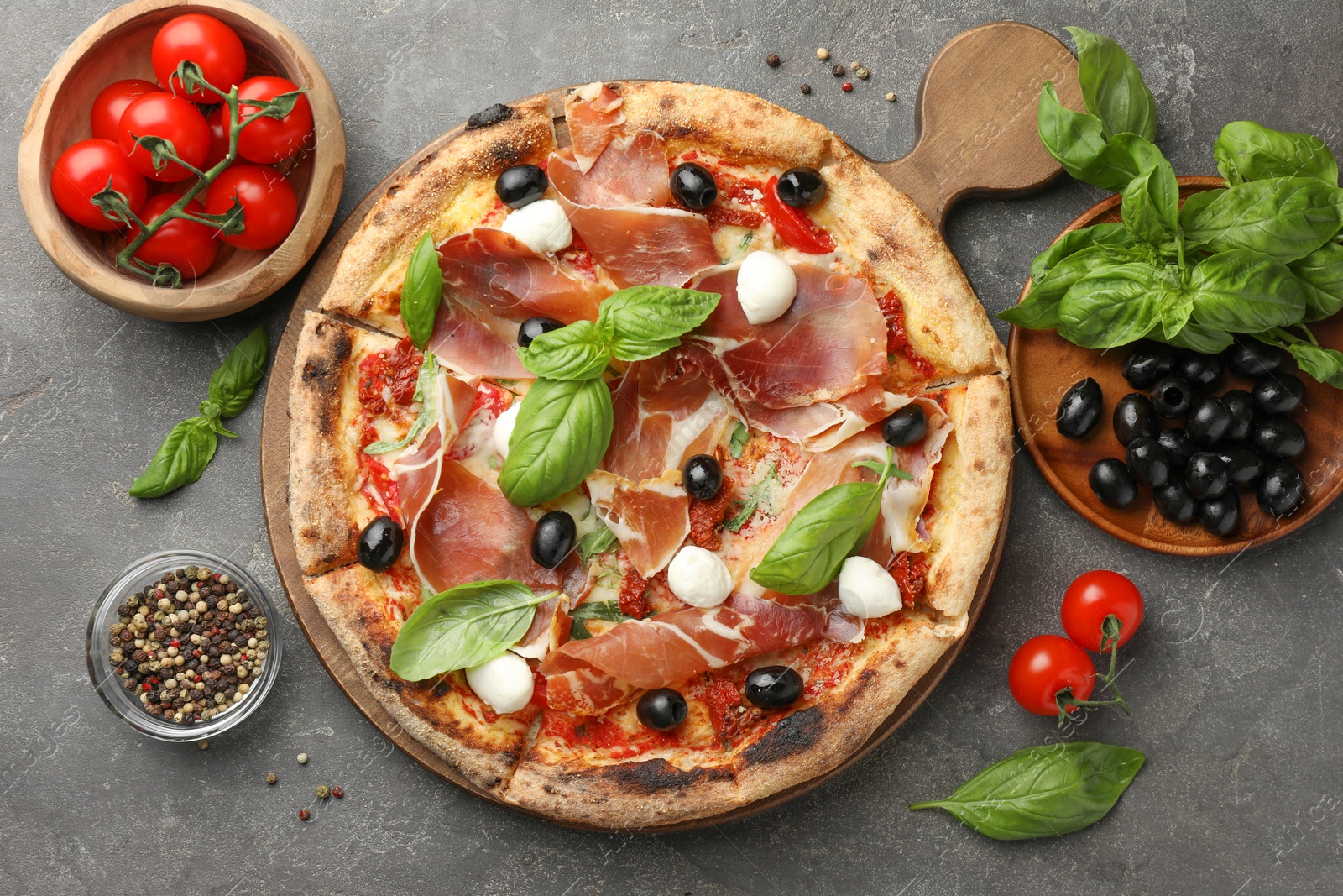 Photo of Tasty pizza with cured ham, olives, mozzarella cheese, tomatoes and arugula on gray table, flat lay