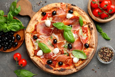 Photo of Tasty pizza with cured ham, olives, mozzarella cheese, tomatoes and arugula on gray table, flat lay