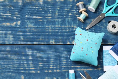 Pincushion with pins and other sewing tools on blue wooden table, flat lay. Space for text