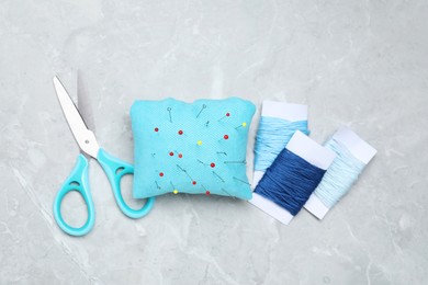 Light blue pincushion with sewing pins, scissors and threads on grey table, flat lay