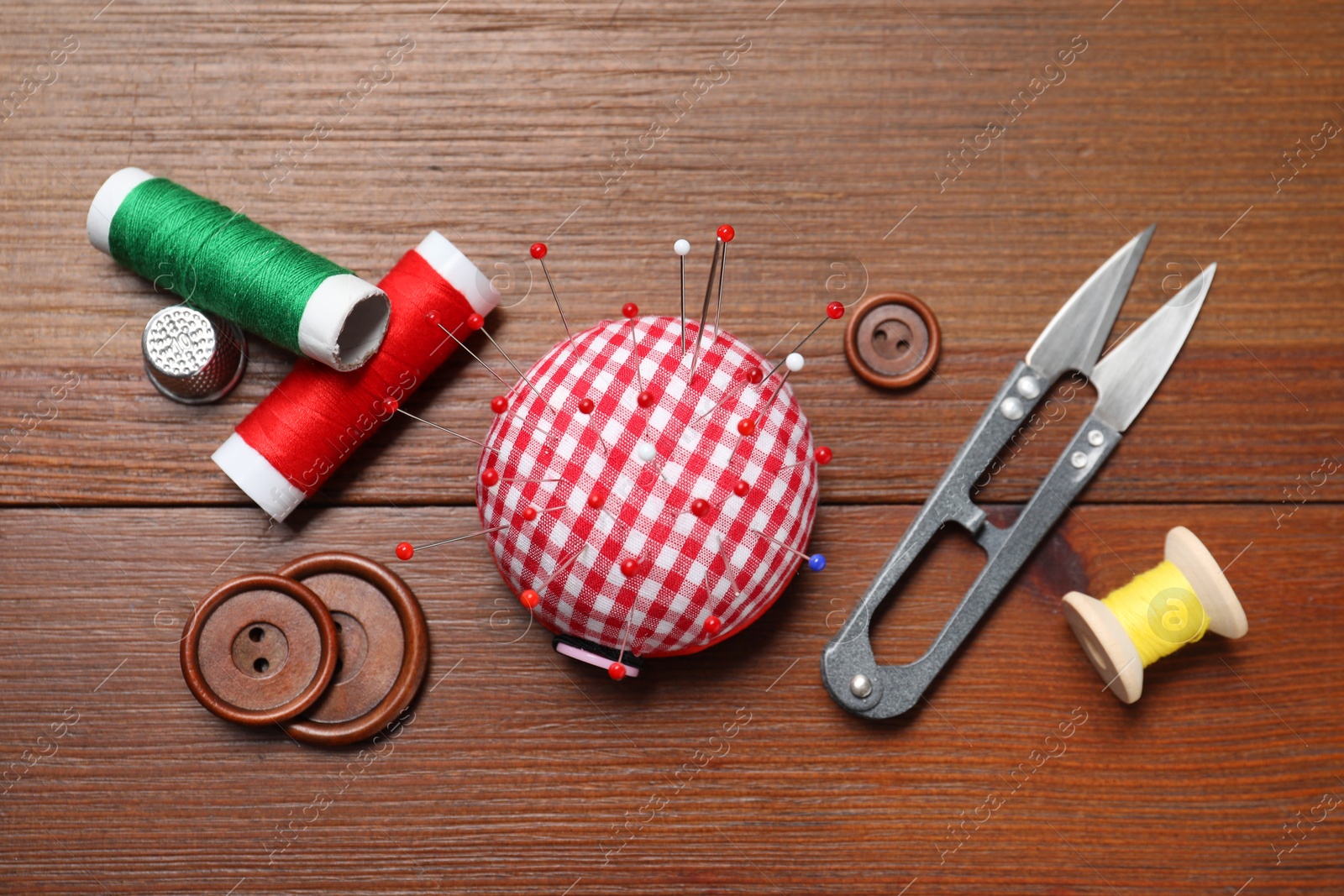 Photo of Checkered pincushion with sewing pins, spools of threads, cutter and buttons on wooden table, flat lay