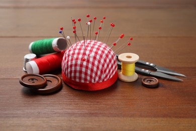 Checkered pincushion with sewing pins, spools of threads, cutter and buttons on wooden table