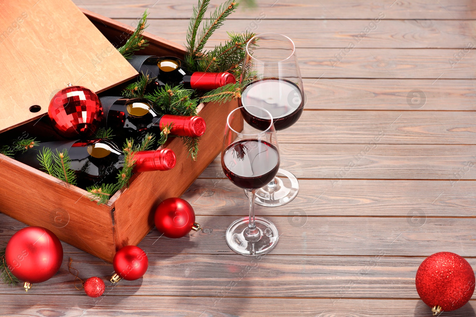 Photo of Wooden crate with bottles of wine, glasses, fir twigs and red Christmas balls on table. Space for text