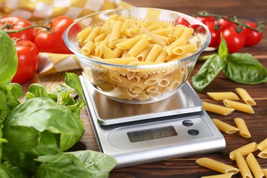 Kitchen scale with bowl of pasta among tomatoes and basil on wooden table, closeup