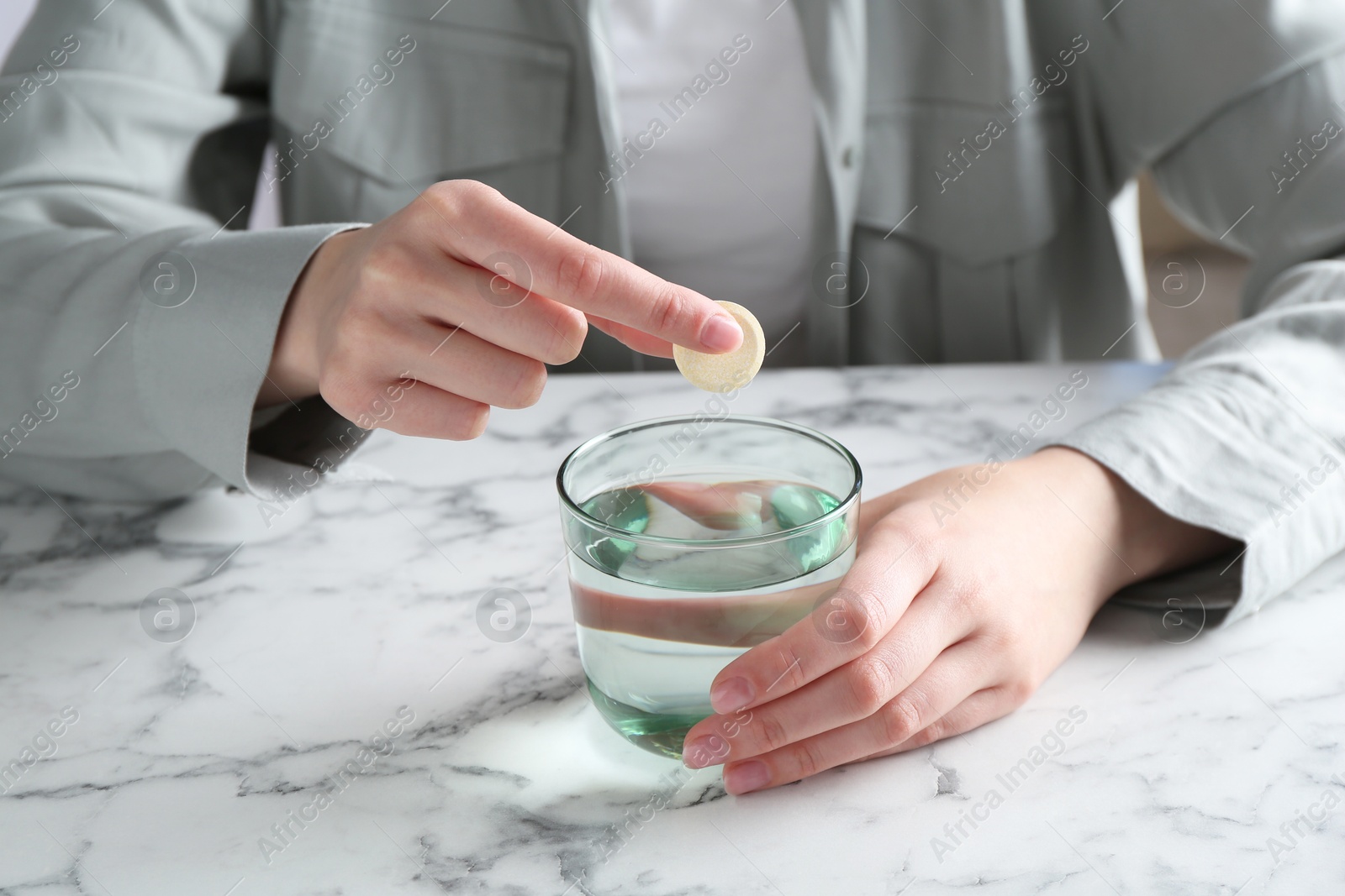 Photo of Woman putting effervescent pill into glass of water at white marble table, closeup