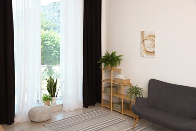 Photo of Shelving unit, sofa, green houseplants, pouffe and elegant curtains in room. Interior design