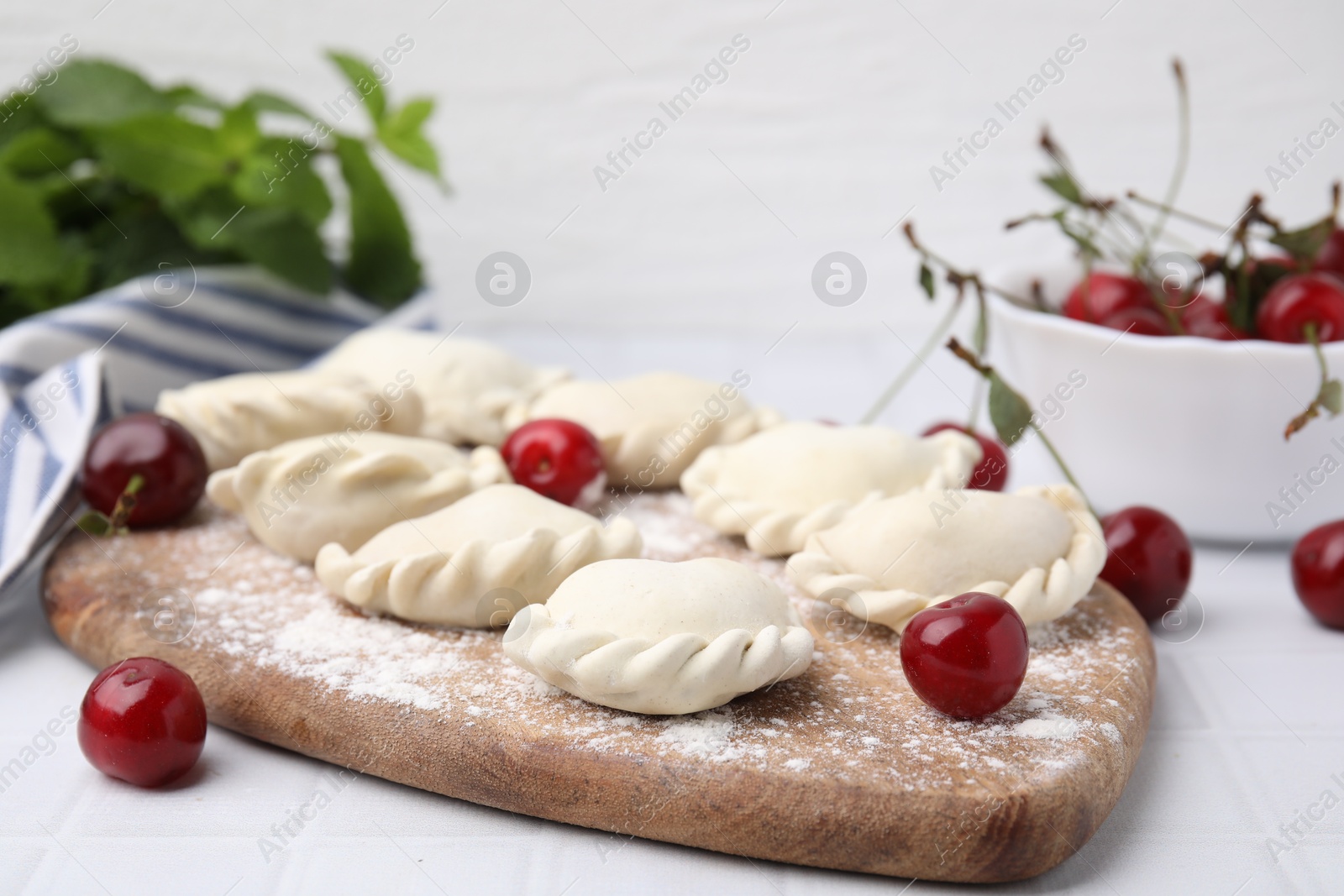 Photo of Raw dumplings (varenyky) and fresh cherries on white tiled table, closeup