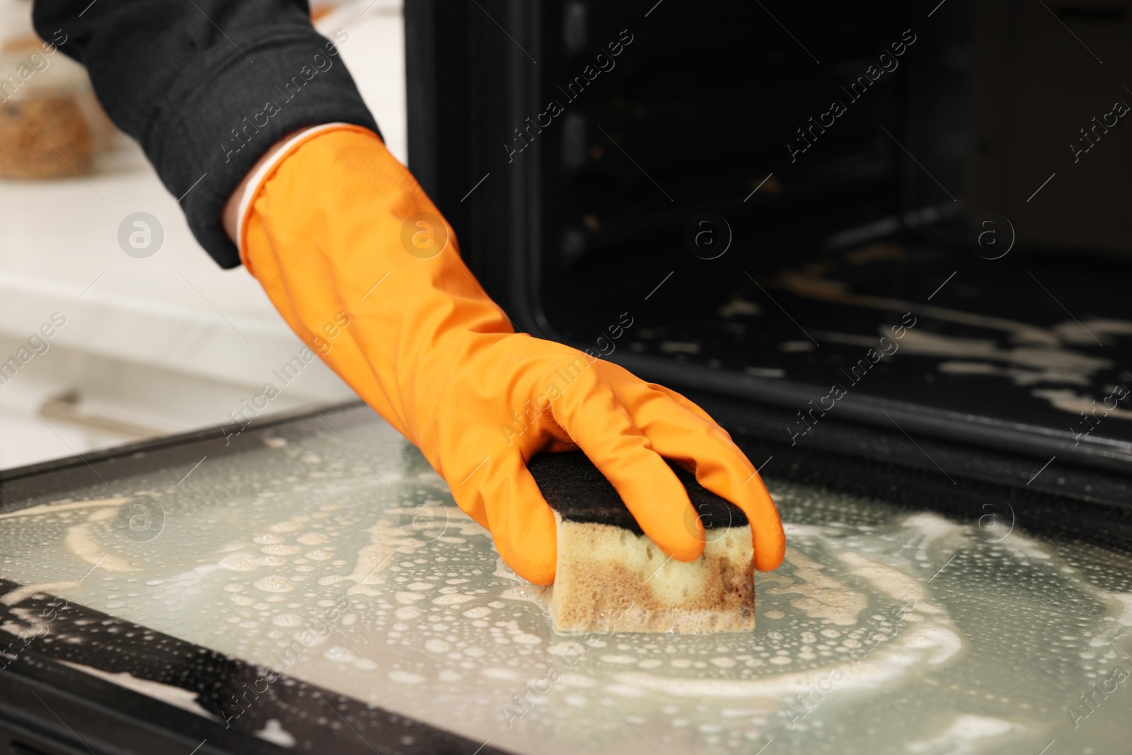 Photo of Woman cleaning oven door with sponge in kitchen, closeup