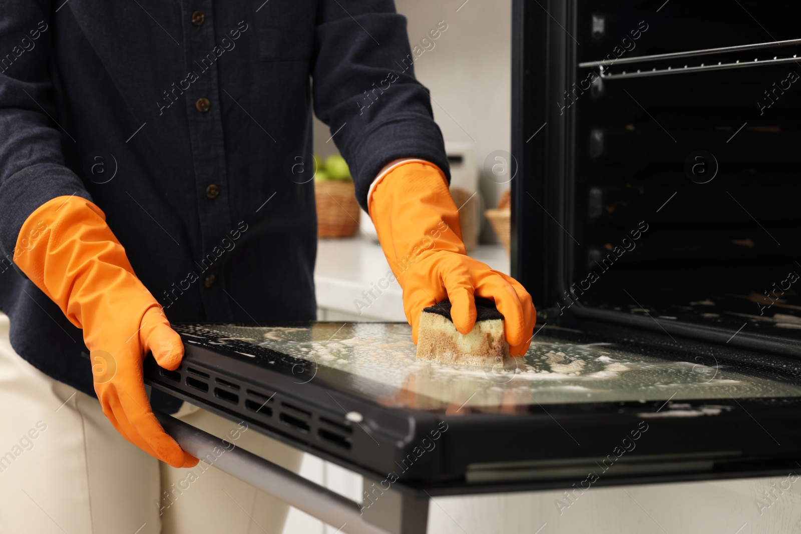 Photo of Woman cleaning oven door with sponge in kitchen, closeup