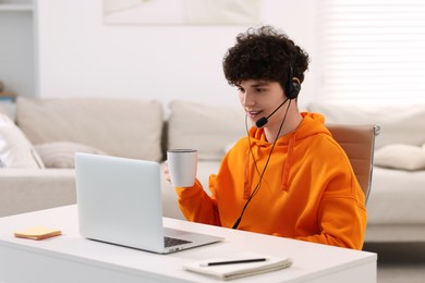 Photo of Teenager in headset holding cup of drink and working with laptop at home. Remote job