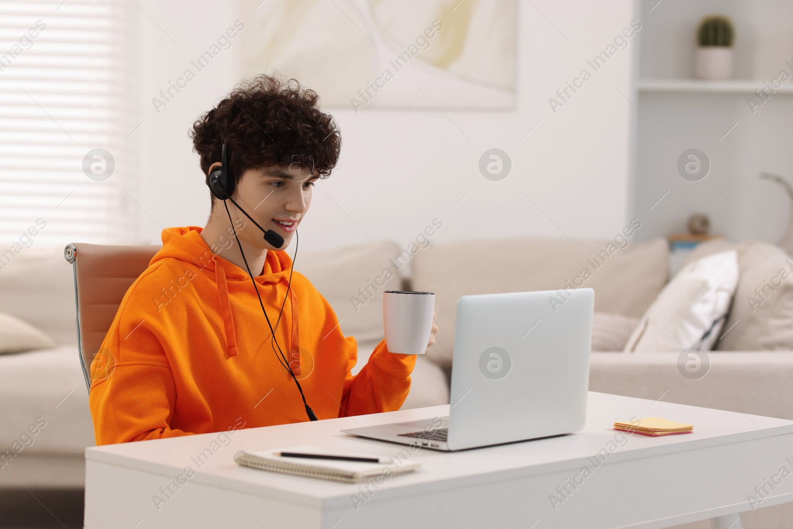 Photo of Teenager in headset holding cup of drink and working with laptop at home. Remote job