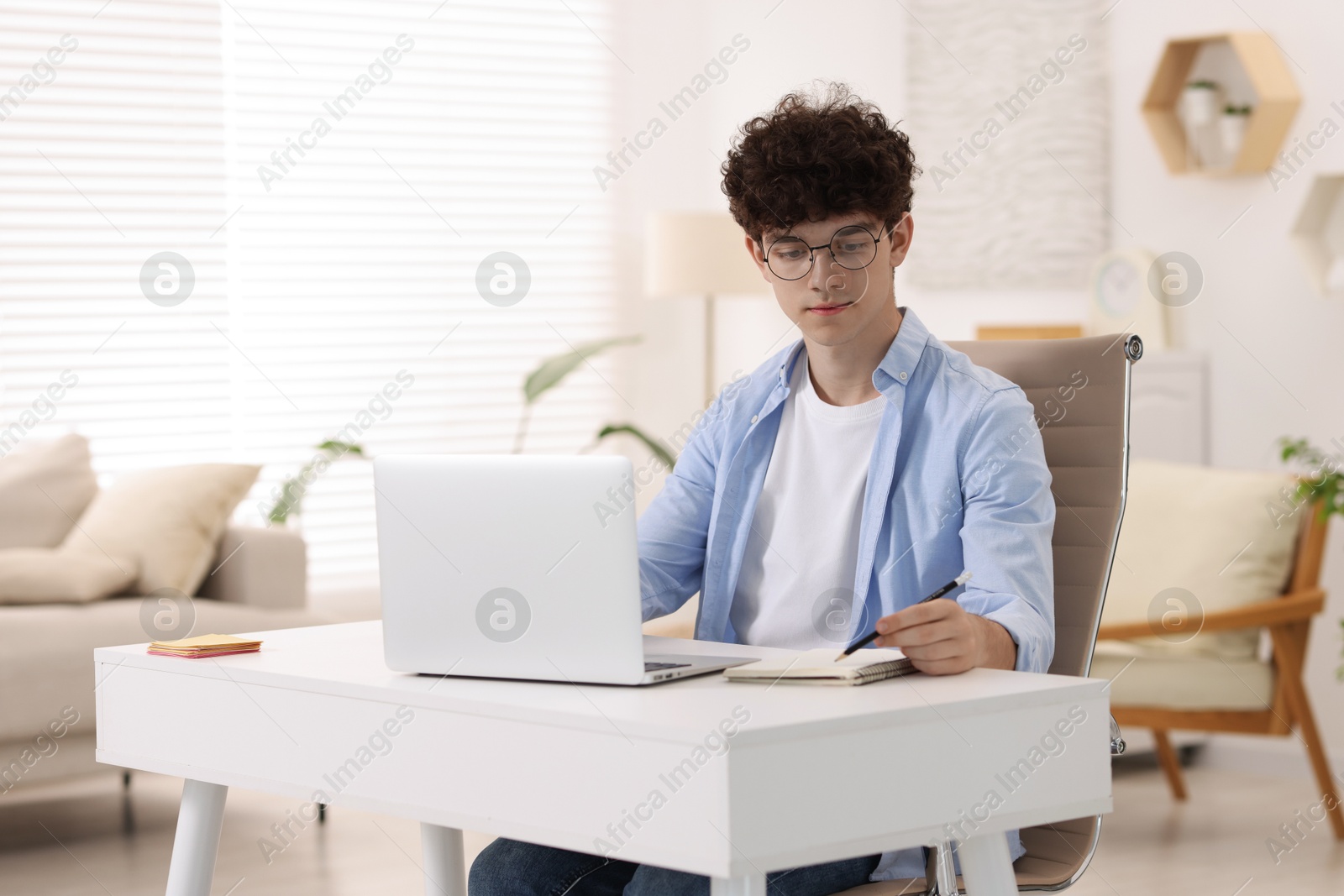 Photo of Teenager taking notes while working with laptop on sofa at home. Remote job