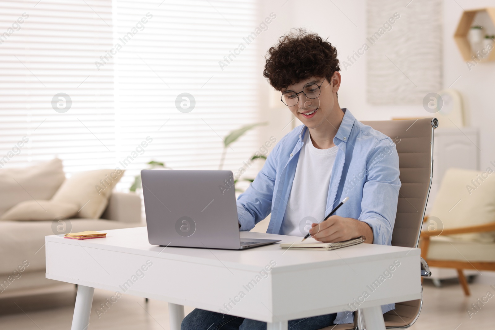 Photo of Teenager taking notes while working with laptop on sofa at home. Remote job