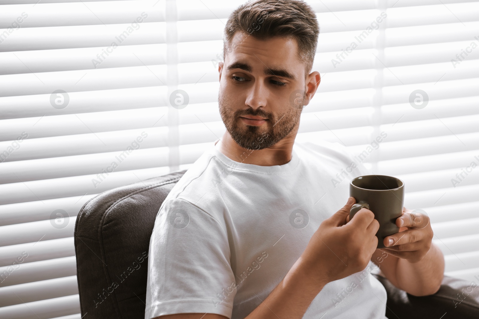 Photo of Man with cup of drink sitting on armchair near window blinds indoors