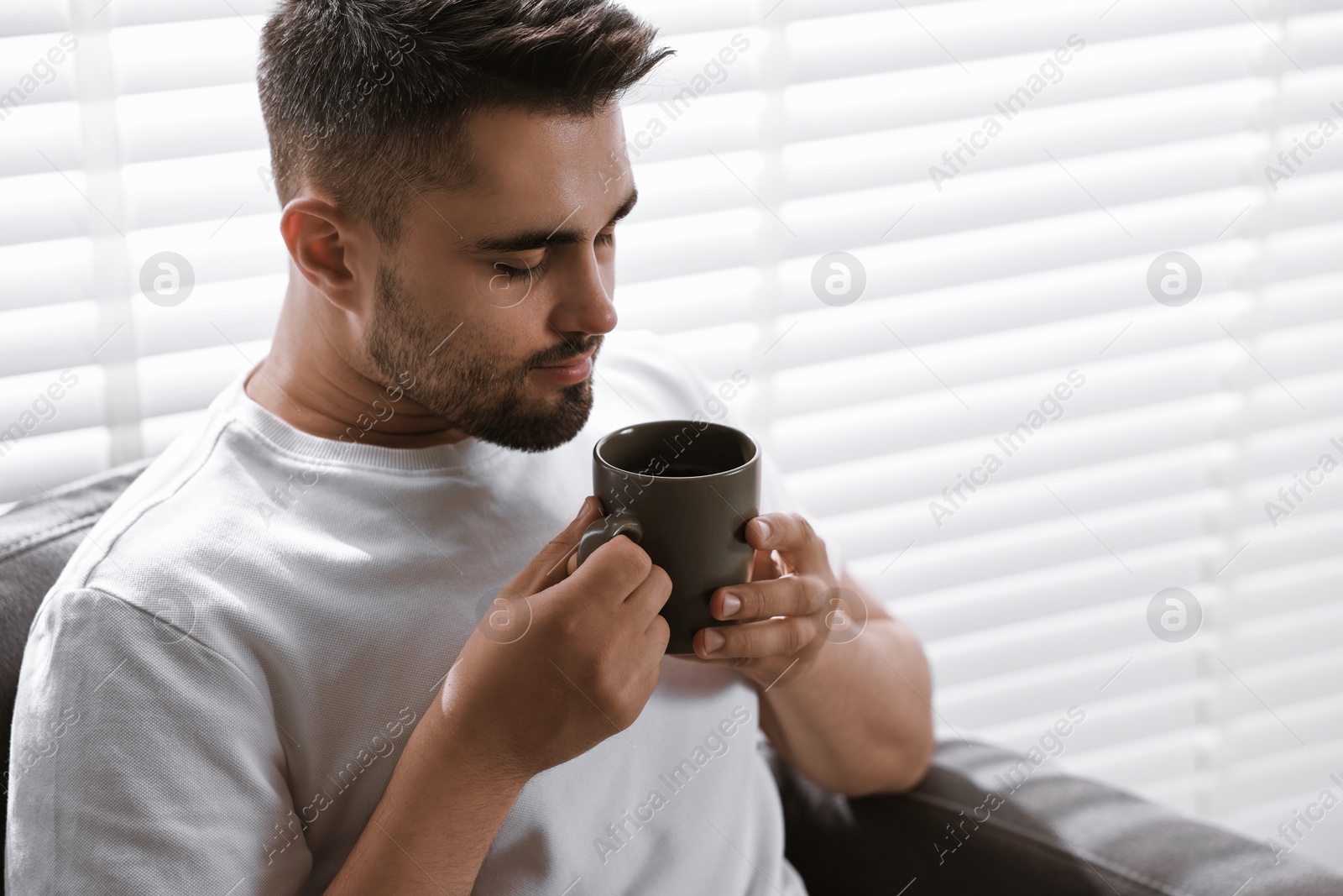 Photo of Man with cup of drink sitting on armchair near window blinds indoors