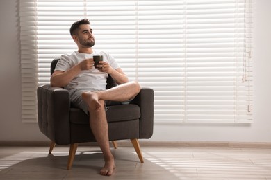 Photo of Man with cup of drink sitting on armchair near window blinds indoors, space for text