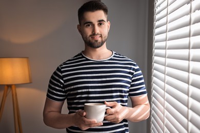Photo of Man with cup of drink near window blinds at home