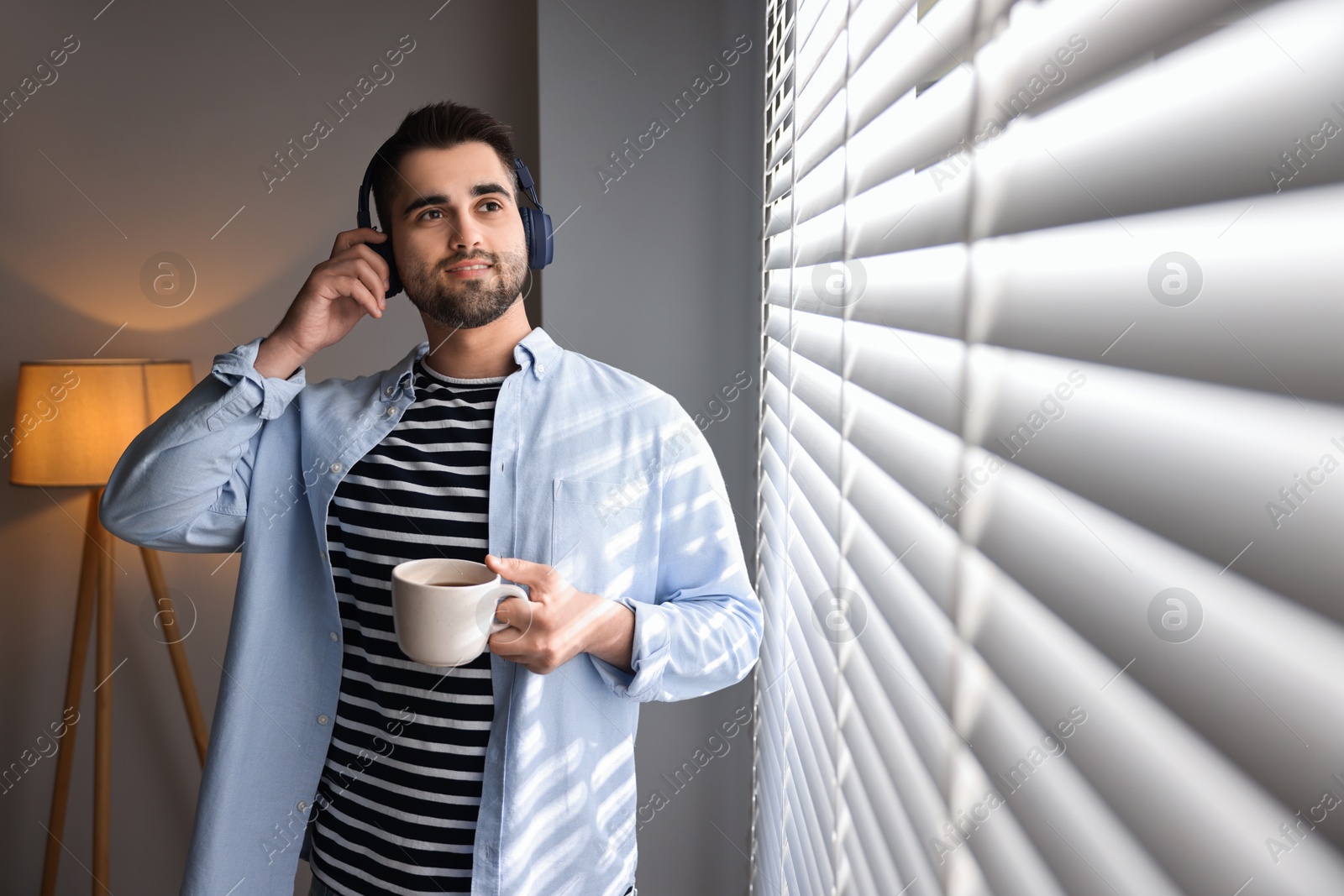 Photo of Man with headphones and cup of drink listening to music near window blinds at home, space for text