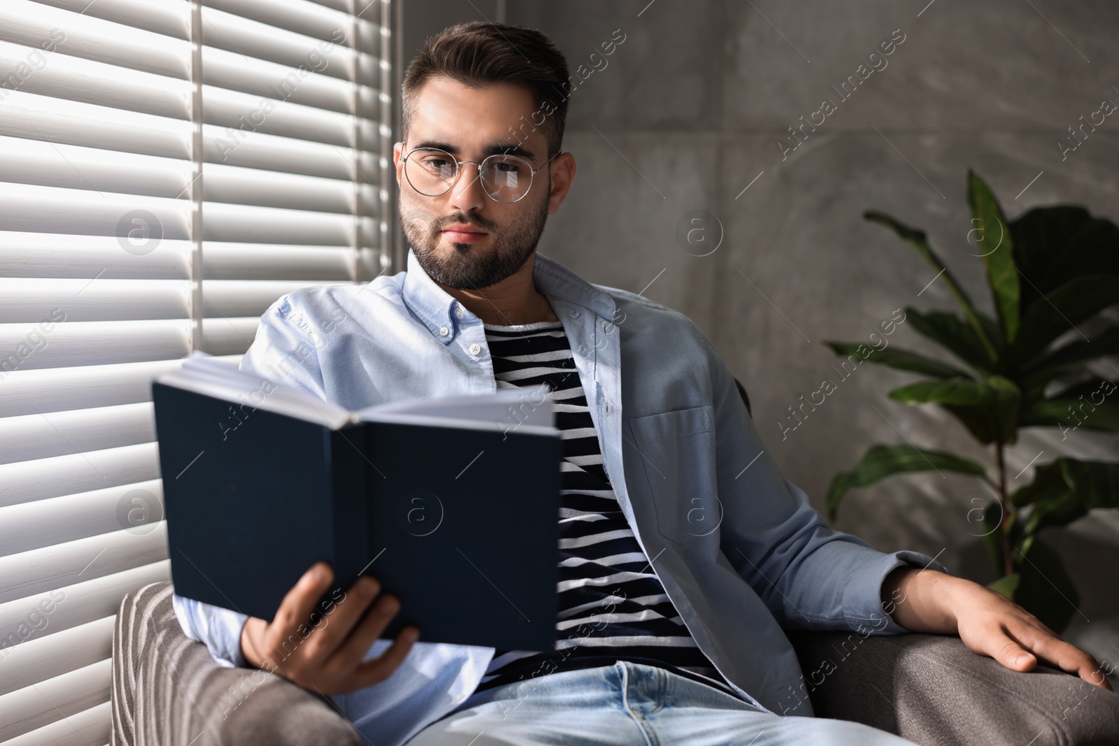 Photo of Man reading book near window blinds indoors