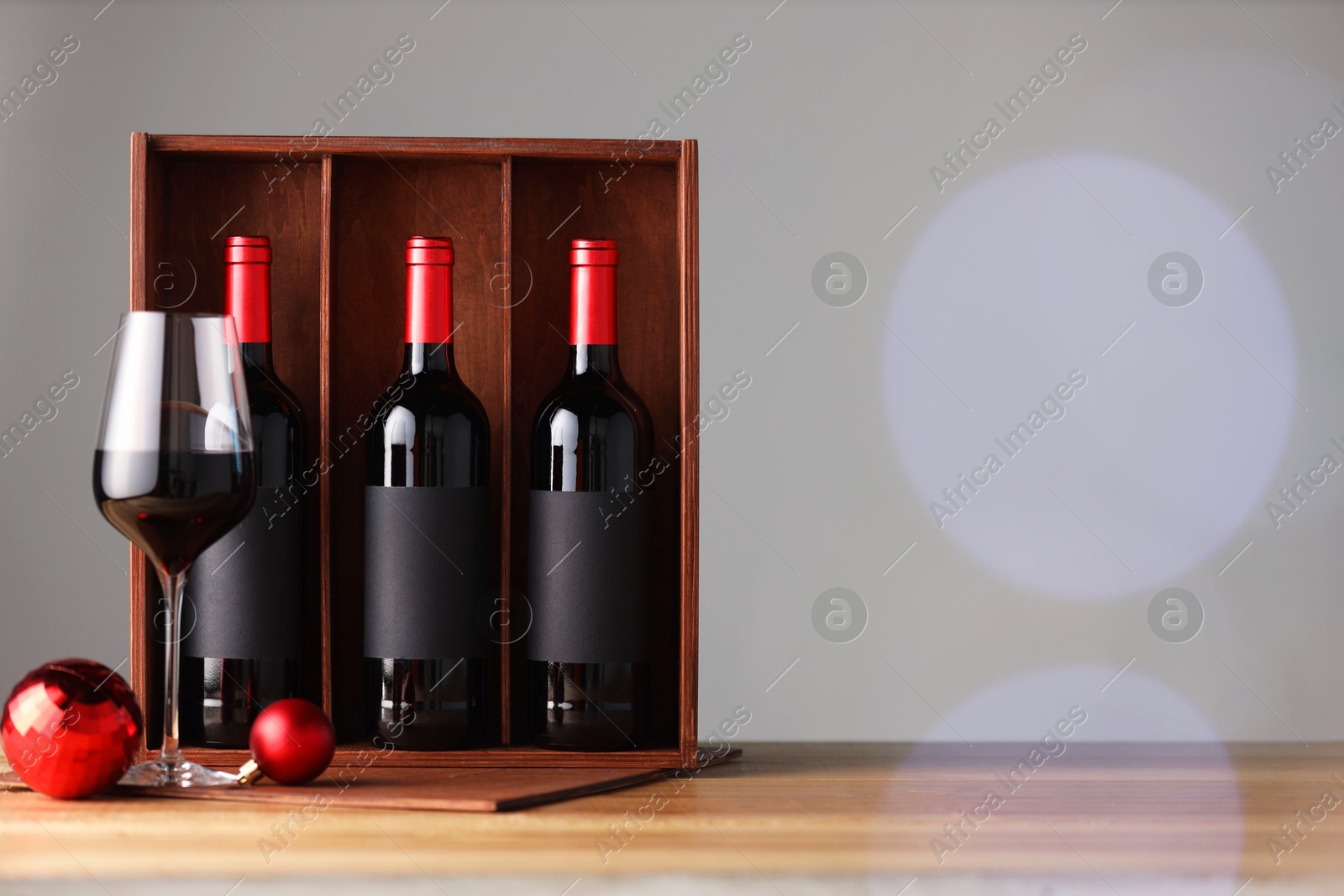 Photo of Wooden gift box with bottles of wine, glasses and red Christmas balls on table, space for text