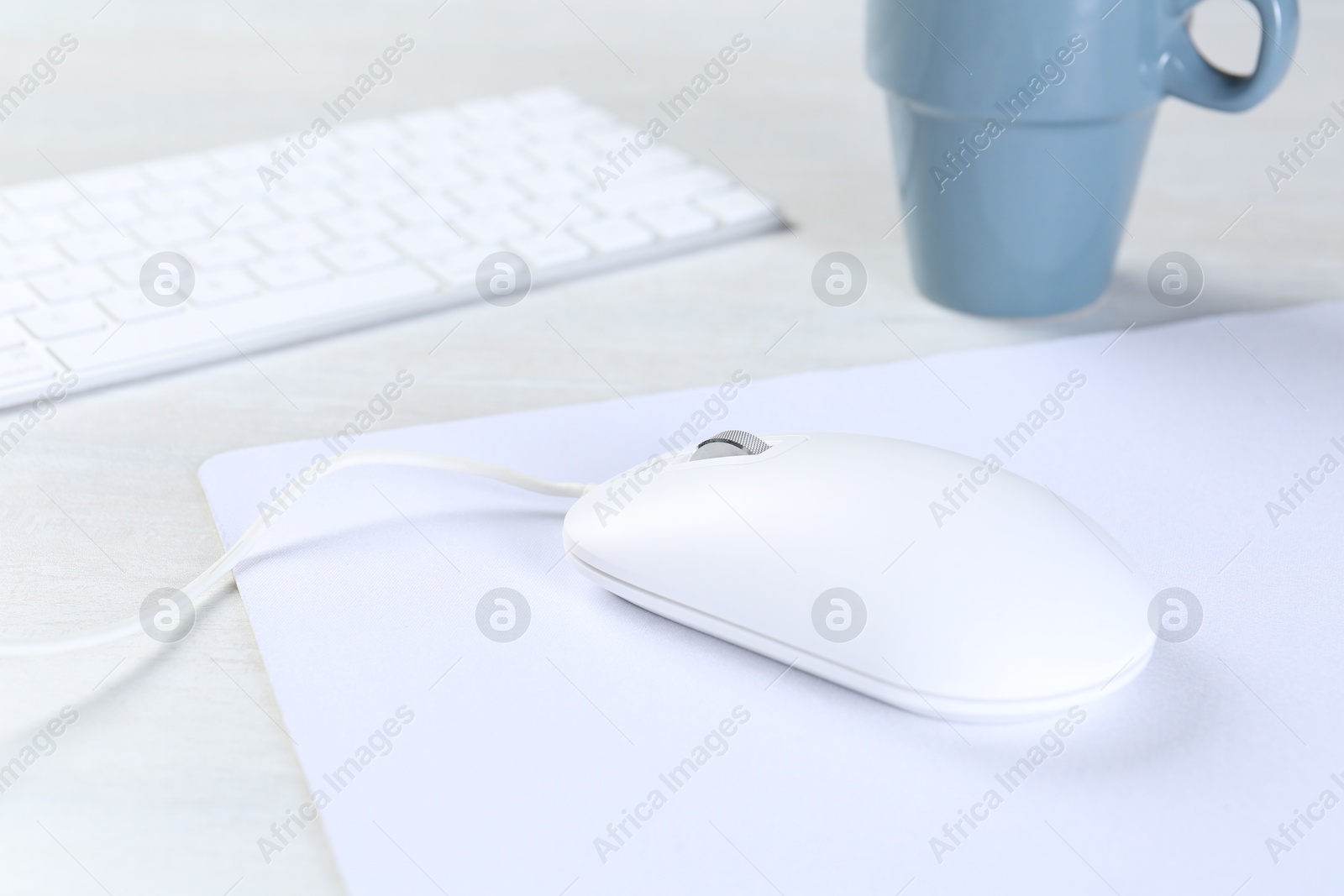 Photo of Computer mouse with mousepad, keyboard and cup on light wooden table, closeup