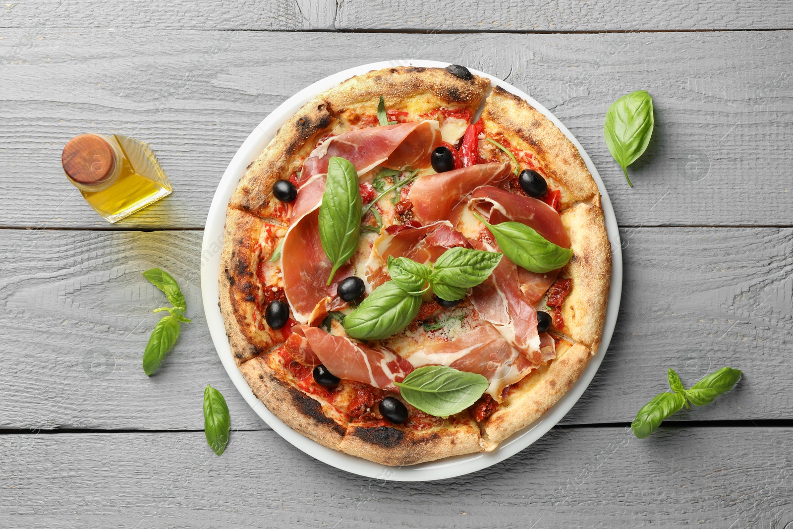 Photo of Tasty pizza with cured ham, olives, sun-dried tomato, basil and oil on gray wooden table, top view