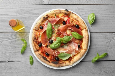 Photo of Tasty pizza with cured ham, olives, sun-dried tomato, basil and oil on gray wooden table, top view