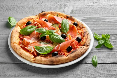 Photo of Tasty pizza with cured ham, olives, sun-dried tomato and basil on gray wooden table, closeup