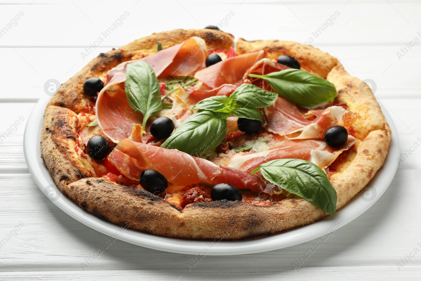 Photo of Tasty pizza with cured ham, olives, sun-dried tomato and basil on white wooden table, closeup