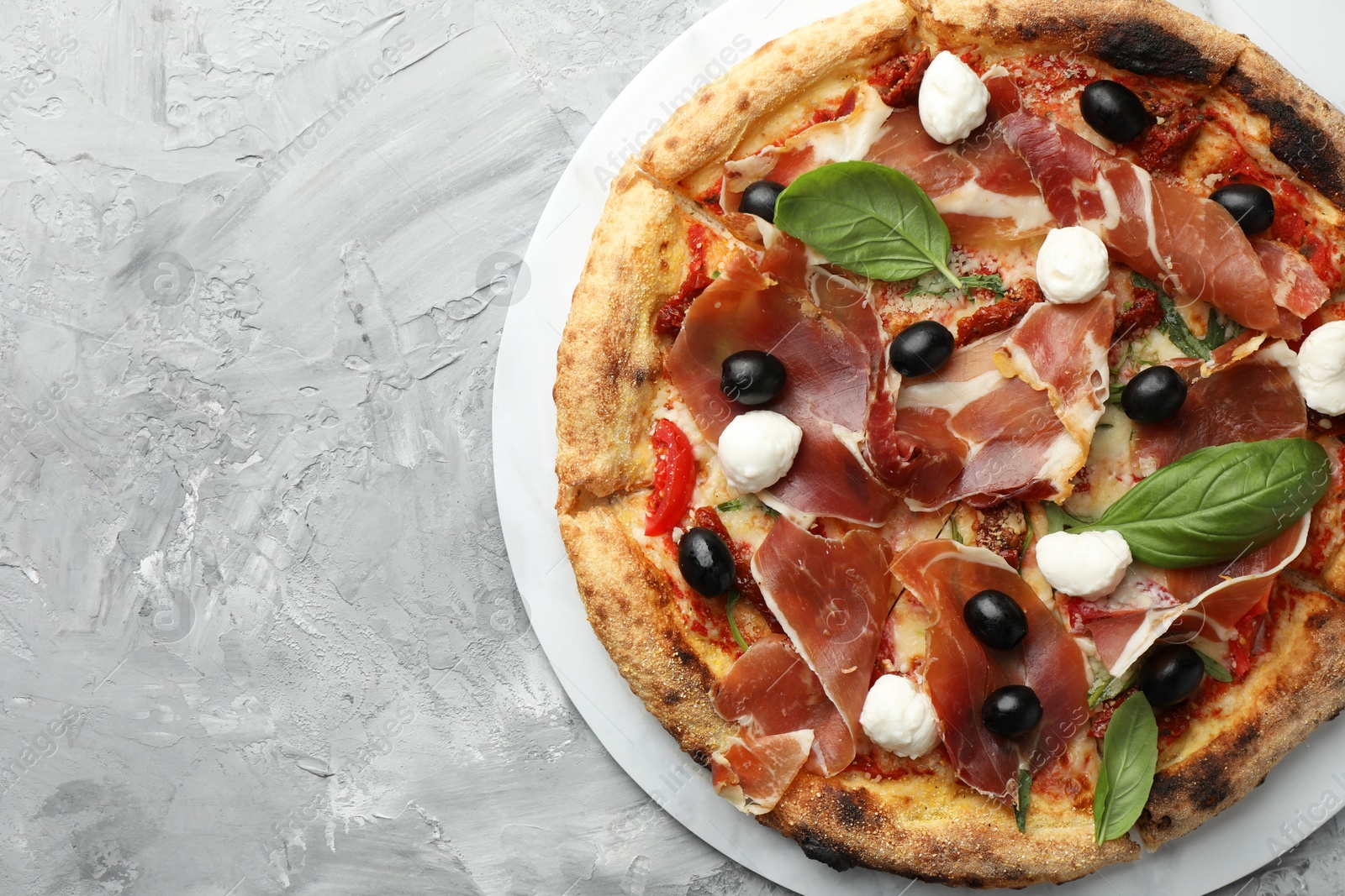 Photo of Tasty pizza with cured ham, olives, sun-dried tomato and basil on gray textured table, top view. Space for text