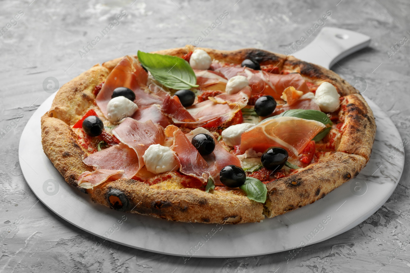 Photo of Tasty pizza with cured ham, olives, mozzarella cheese, sun-dried tomato and basil on gray textured table