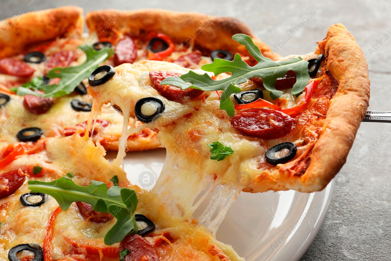 Photo of Taking piece of tasty pizza with smoked sausages, olives, pepper and parsley at gray table, closeup