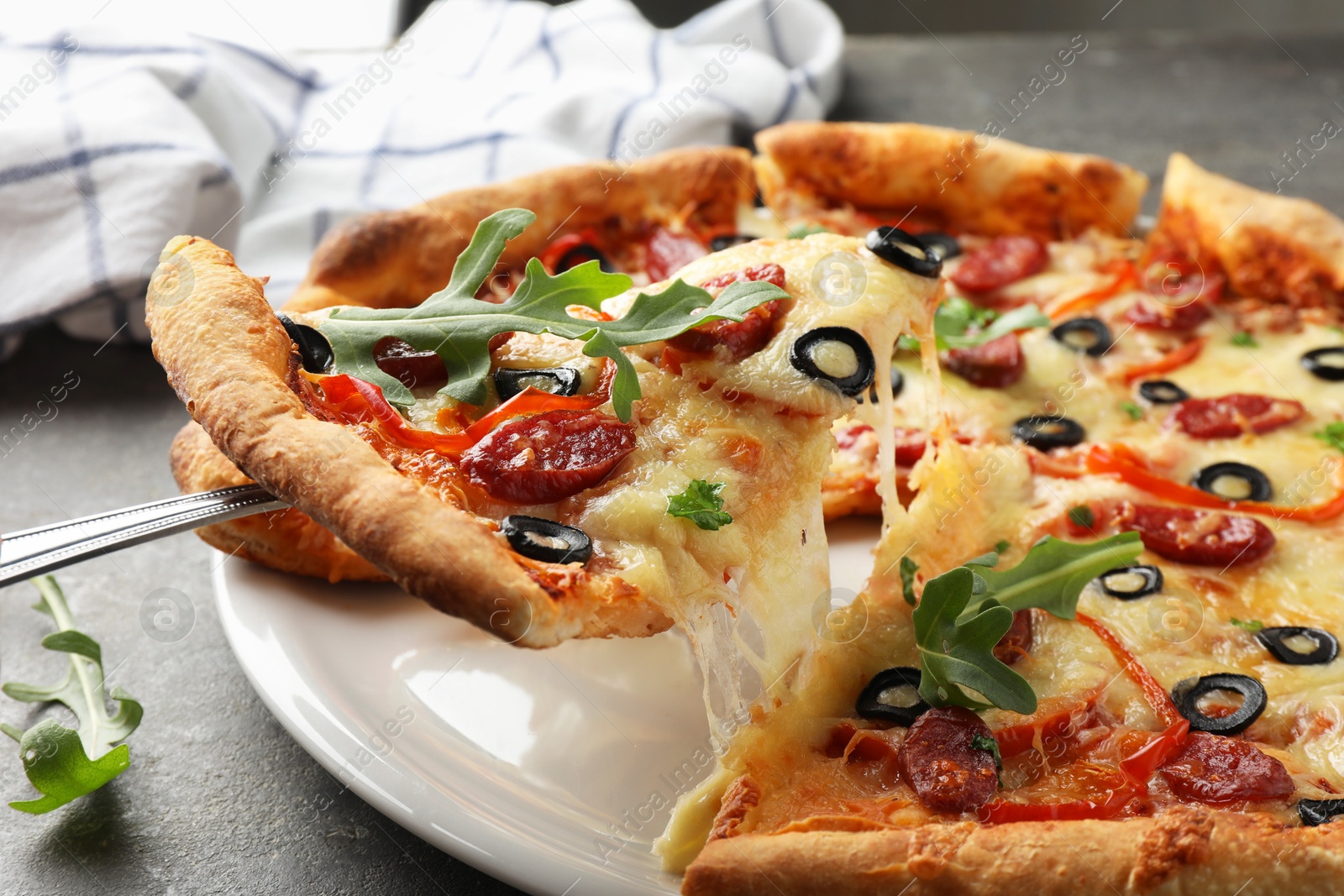 Photo of Taking piece of tasty pizza with smoked sausages, olives, pepper and parsley at gray table, closeup