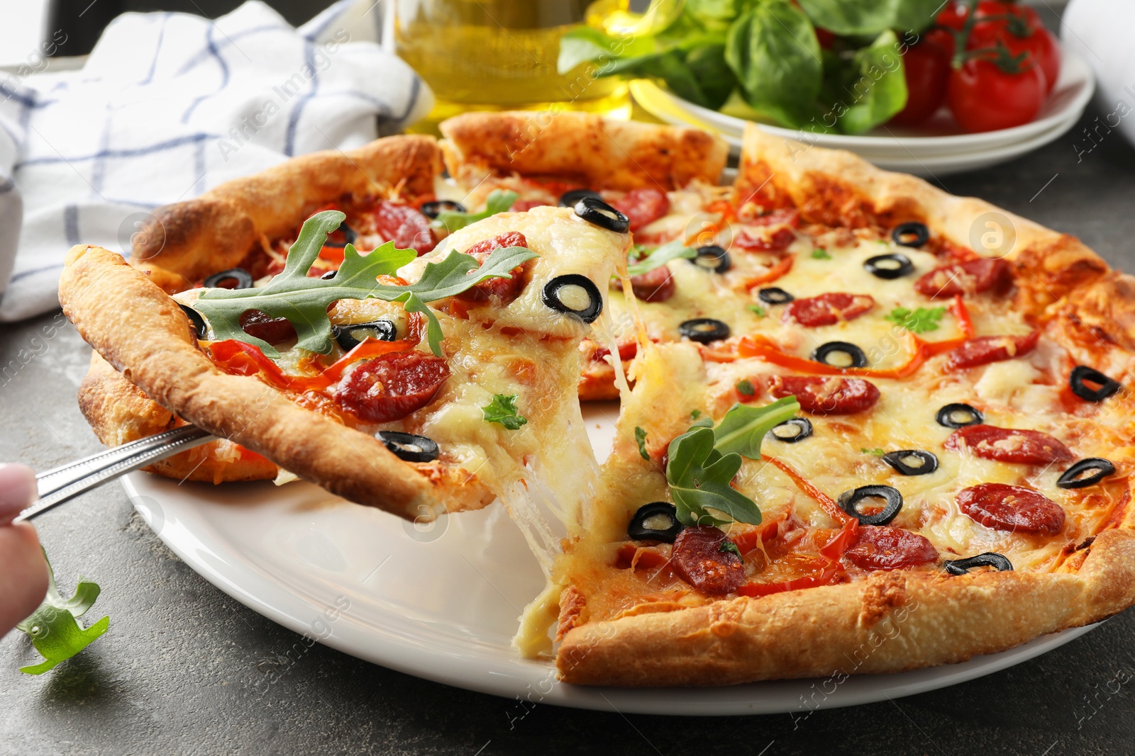 Photo of Taking piece of tasty pizza with smoked sausages, olives, pepper and parsley at gray table, closeup