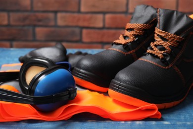 Photo of Pair of working boots, reflective vest and earmuffs on blue wooden surface, closeup