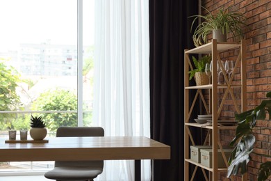 Photo of Shelving unit, table, chair, green houseplants and elegant curtain in room. Interior design