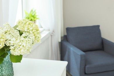 Photo of Beautiful flowers in vase on table near blue armchair indoors, selective focus. Space for text