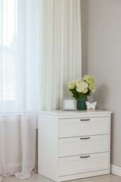 Photo of Chest of drawers, flowers, decorative elements and window with curtains indoors