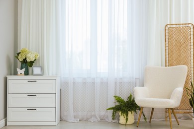 Photo of Soft armchair, chest of drawers, houseplant and window with curtains in room. Interior design